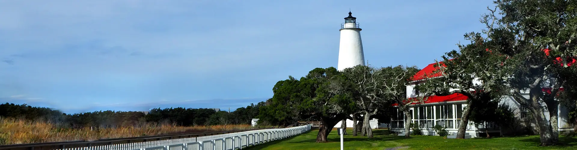 The short white Ocracoke Lighthouse above a mown green lawn, its red-roofed keeper's house and white picket fence beside it