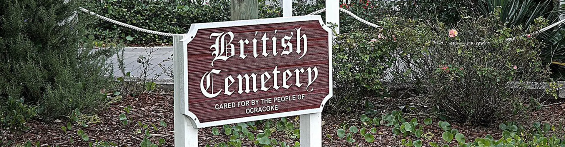 A carved wooden sign reading British Cemetery, cared for by the people of Ocracoke, standing among low shrubs