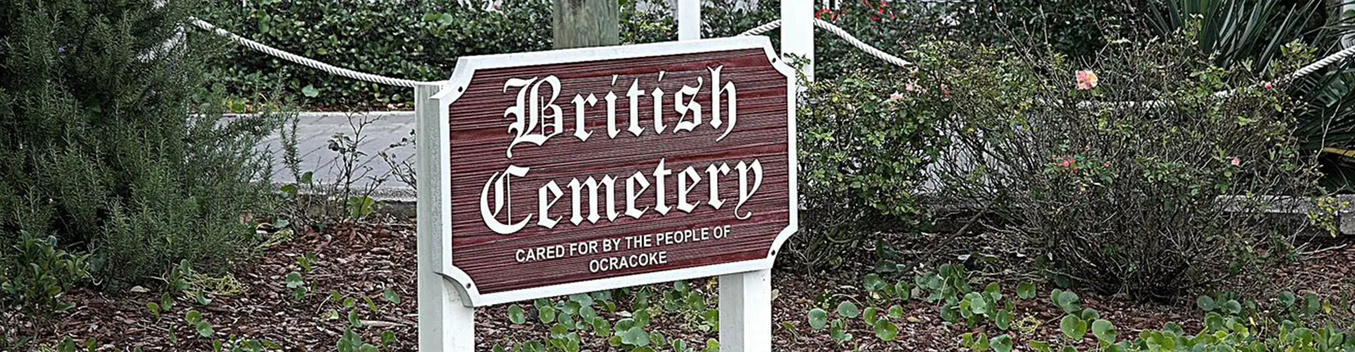 A carved wooden sign reading British Cemetery, cared for by the people of Ocracoke, standing among low shrubs