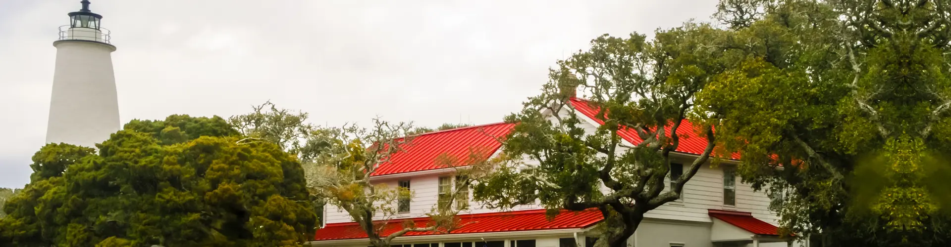 The white tower of Ocracoke Lighthouse rising past live oaks beside its red-roofed keeper's house