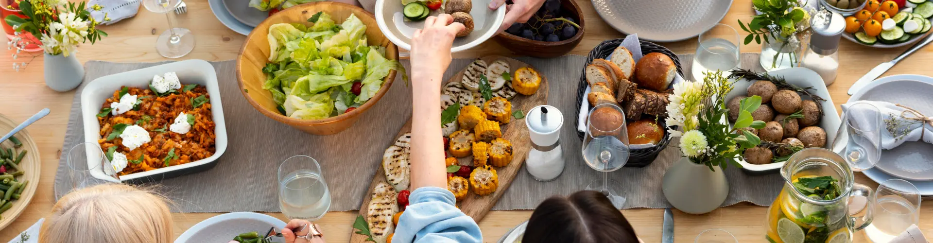 An overhead view of a table crowded with shared dishes, salads and glasses, hands reaching in from every side