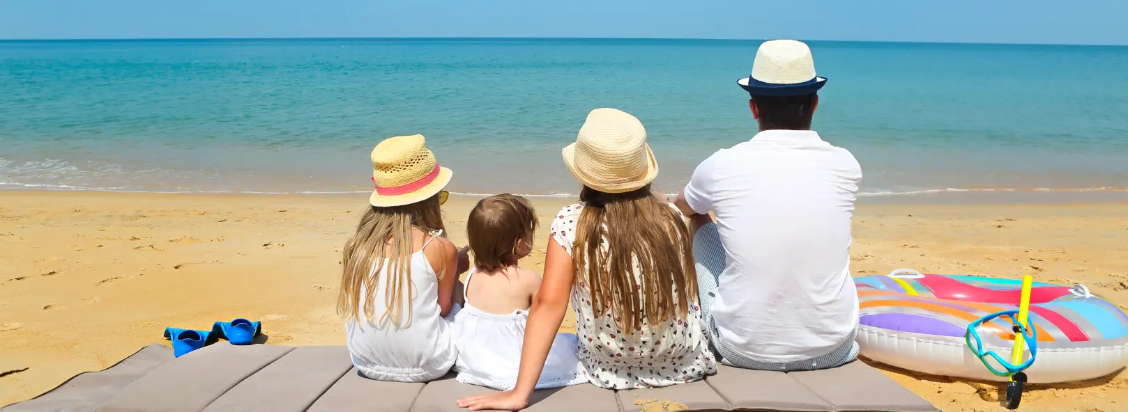 A family of four in straw hats sitting on a boardwalk with snorkel and rubber ring, looking out at turquoise sea