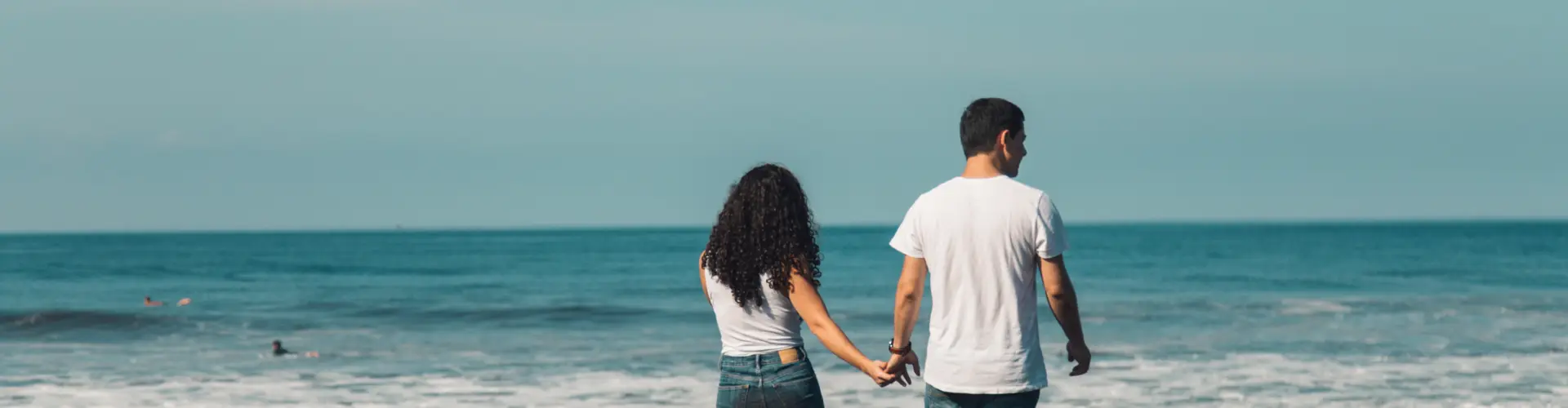 A young couple seen from behind, hand in hand, walking towards a calm grey-green sea