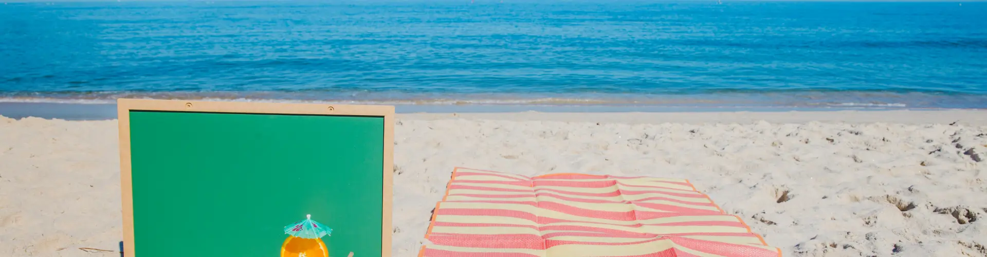 A blank green chalkboard propped in white sand beside a pink striped beach mat and a small cocktail with a paper umbrella