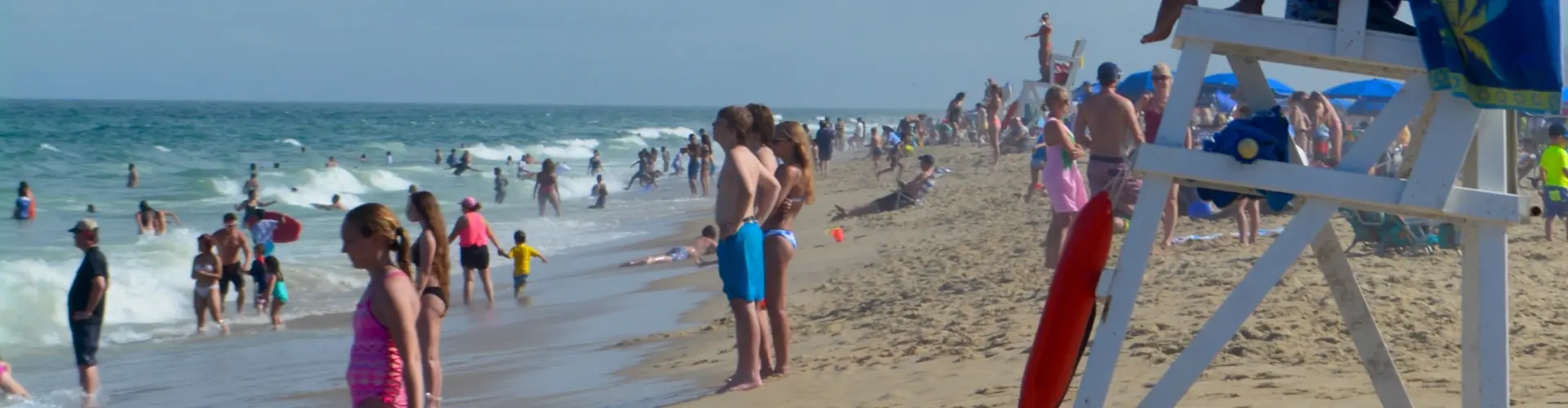 A crowded summer beach of swimmers and umbrellas with a white lifeguard stand in the foreground