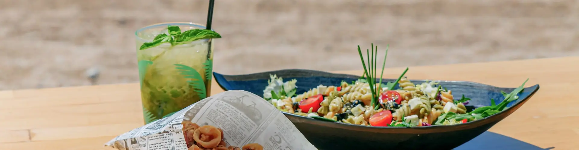 A bowl of fresh salad and a tall mint cocktail set on a table in the sand