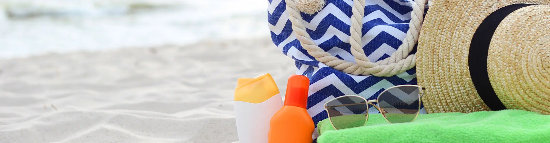 A straw hat, striped beach bag, sunglasses and bottles of sun cream arranged on pale sand