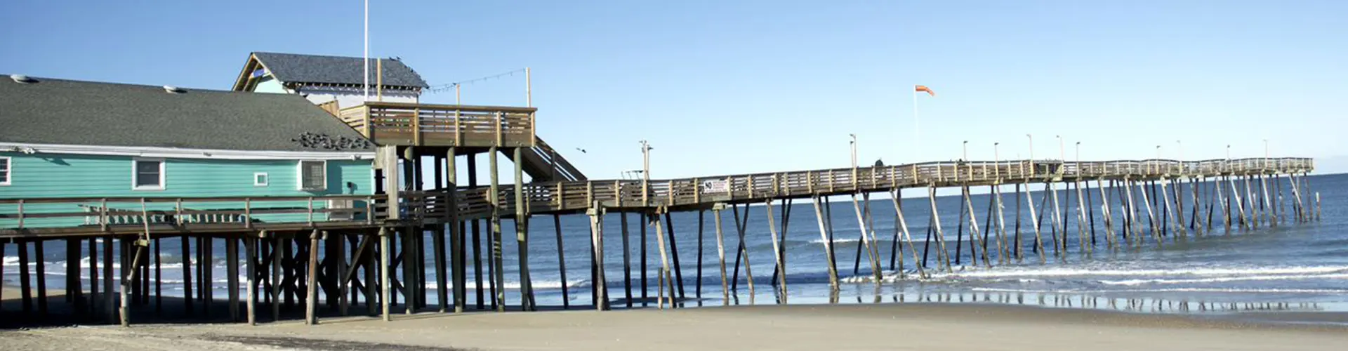 A long timber fishing pier with a pale green pier house on pilings, running out under a clear blue sky