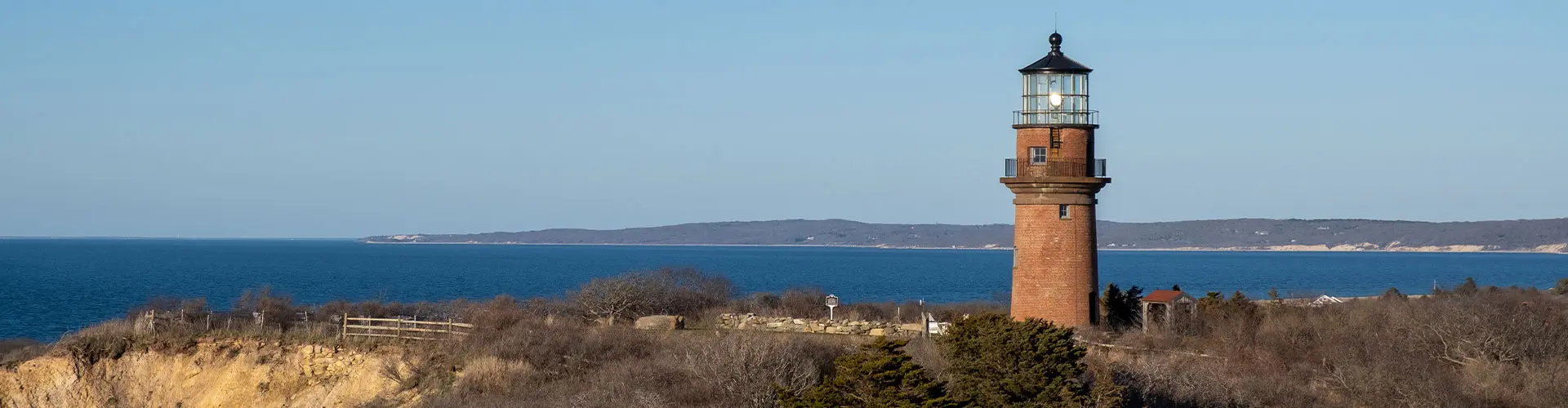 A red-brick lighthouse on a low bluff above deep blue water, winter scrub around it and a distant headland beyond