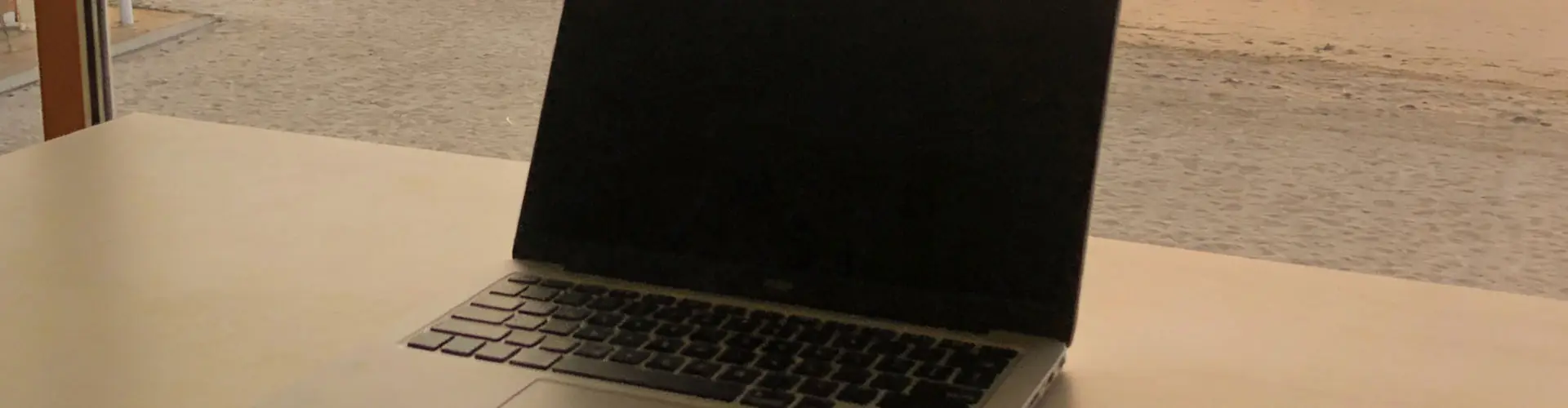 An open laptop with a blank screen on a pale table, a sunlit terrace and sand visible through the window behind