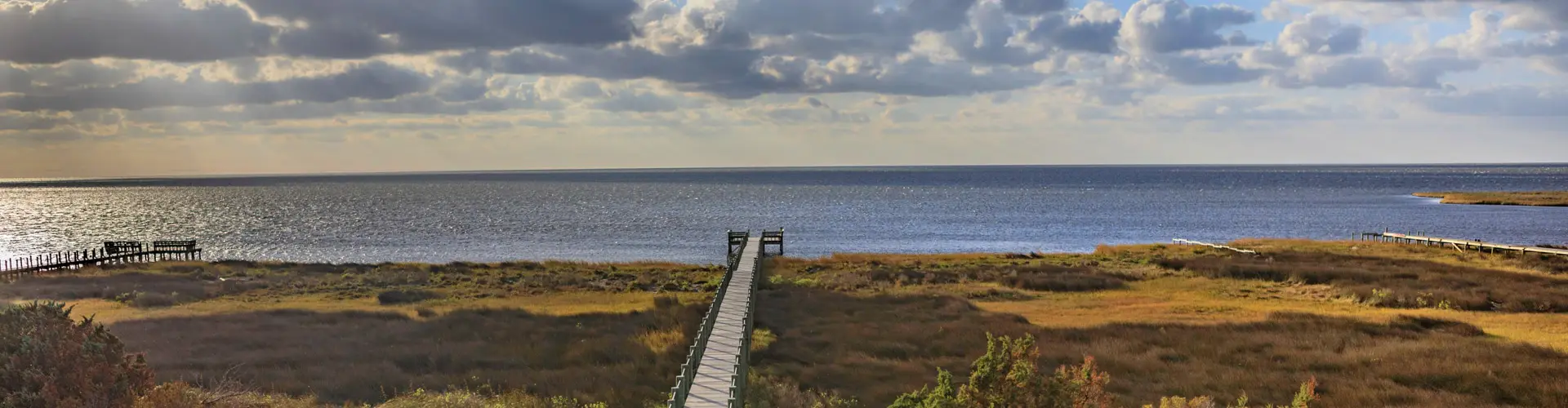 A narrow boardwalk crossing golden marsh grass to a wide, flat sound under a streaked evening sky