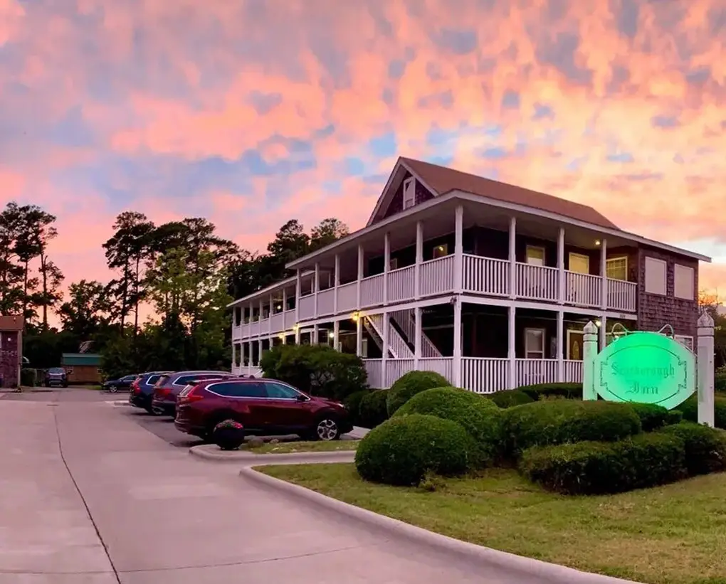 Scarborough Inn under a pink sunset, its two tiers of white porches behind clipped hedges and the illuminated inn sign