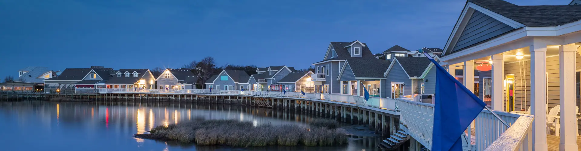 A waterfront row of grey-shingled shops and cottages lit at blue hour along a curving boardwalk, reflections in still water