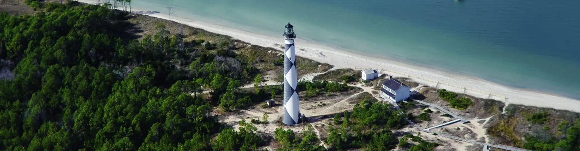 An aerial view along a wooded spit to the diamond-patterned Cape Lookout Lighthouse, beach and green sea to the right