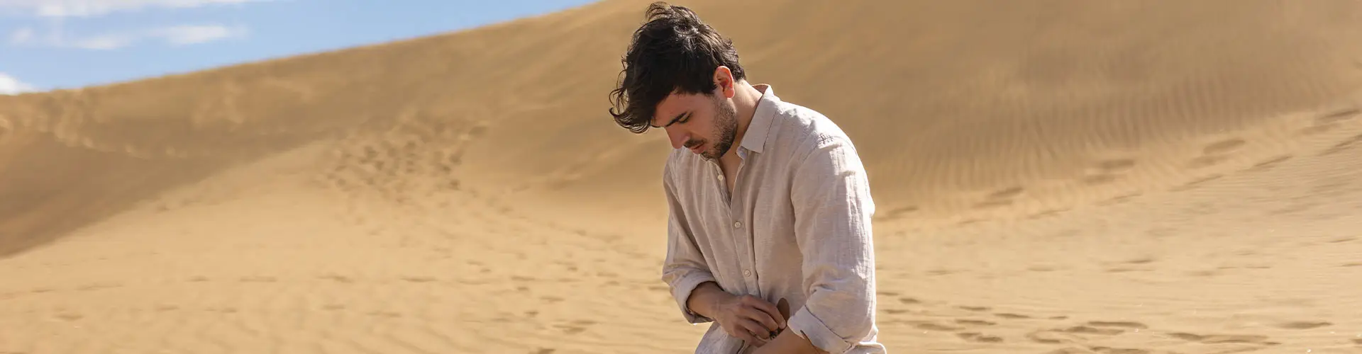 A man crouching on a vast bare sand dune, running the sand through his fingers