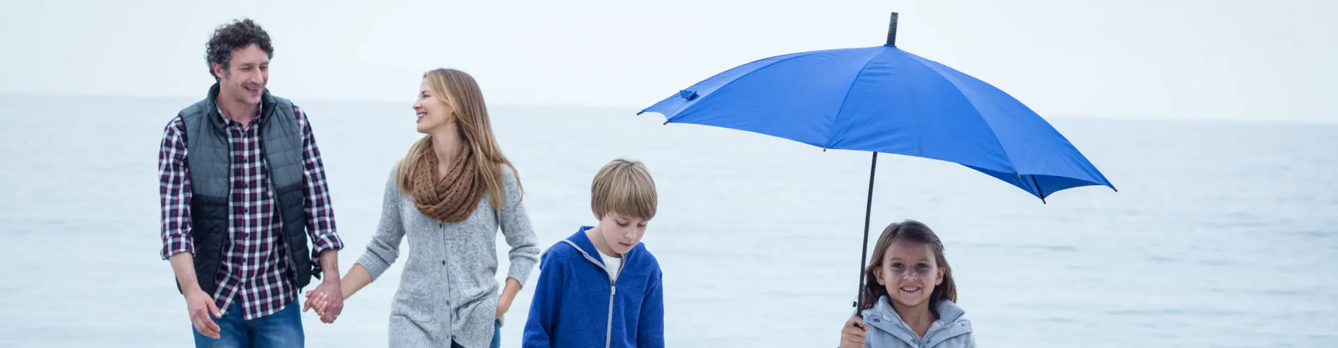A family of four walking the beach in the rain under a large blue umbrella