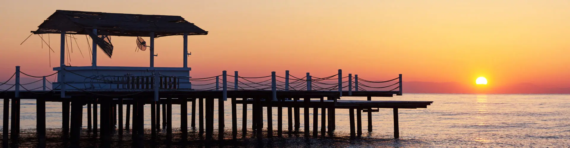 A wooden pier with a covered pavilion running out into a flat sea as the sun sets behind it
