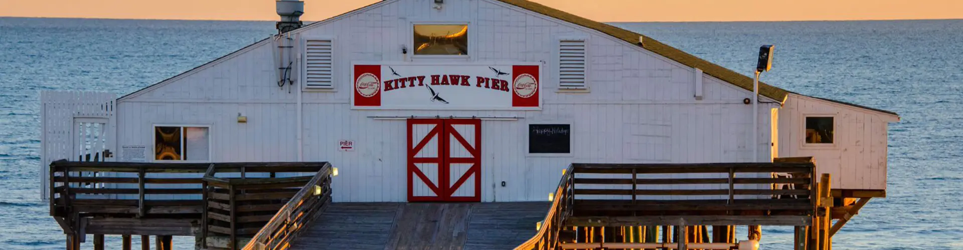 The white clapboard Kitty Hawk Pier house with its red doors, standing over the beach on pilings