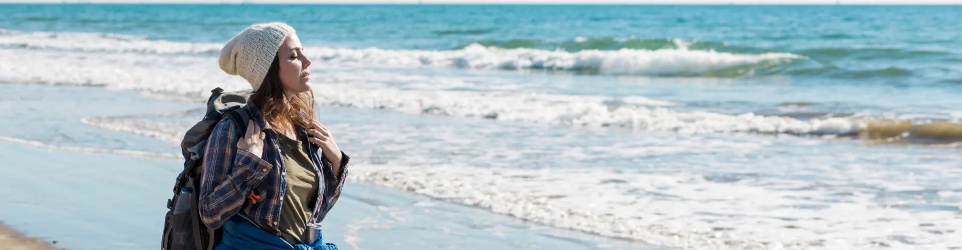 A woman in a knitted hat and rucksack looking out over pale green surf on an empty beach