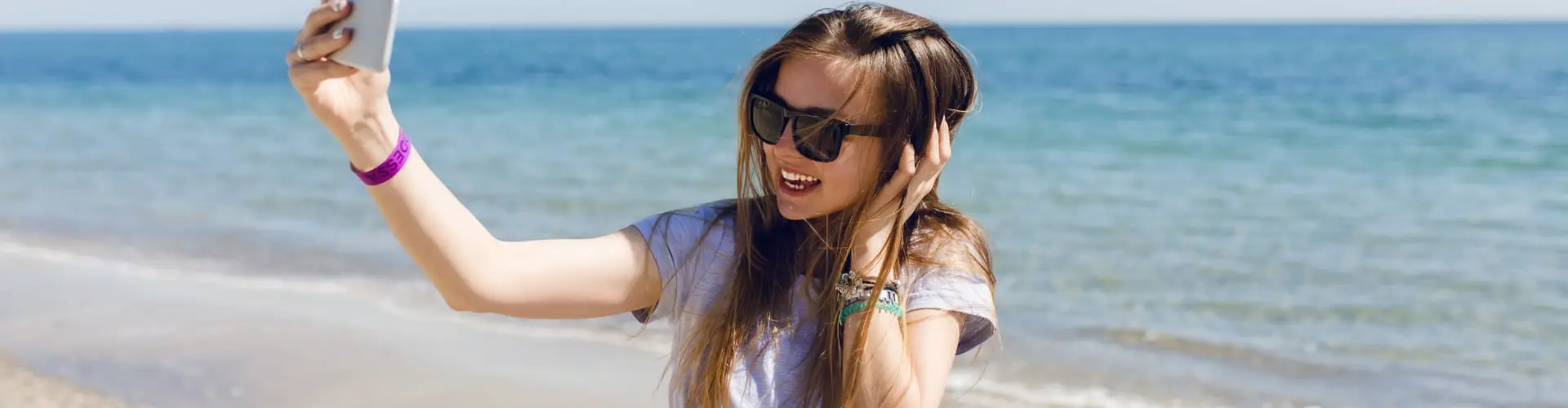 A young woman in sunglasses taking a selfie on the beach, sea behind her