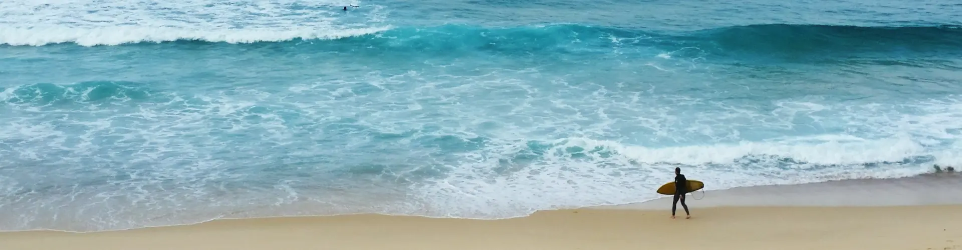 A lone surfer walking out of turquoise surf onto an empty stretch of sand