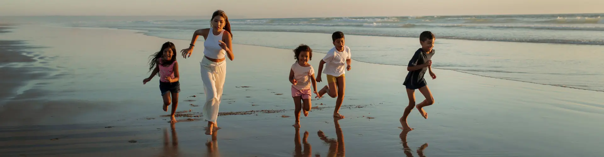 Three children running along wet sand with a woman, their reflections under them