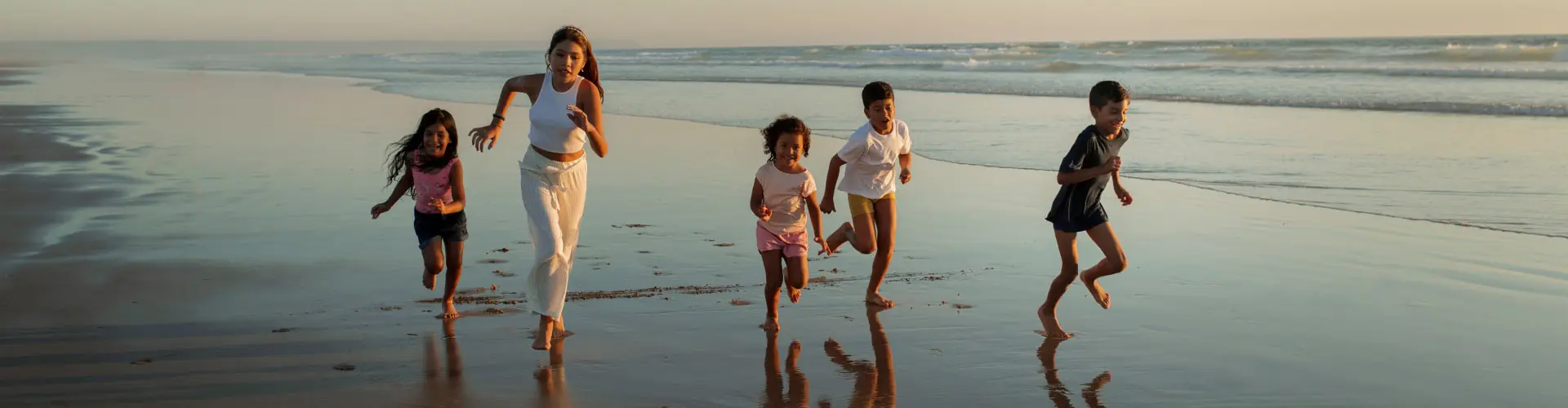 Three children running along wet sand with a woman, their reflections under them
