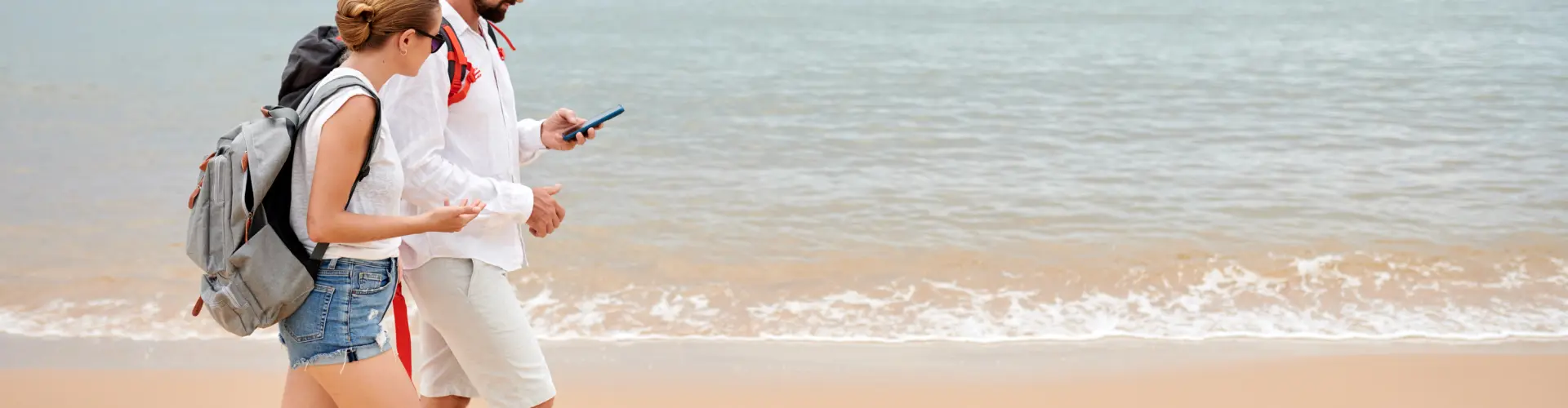 A woman with a rucksack walking barefoot along the tideline, shoes in hand