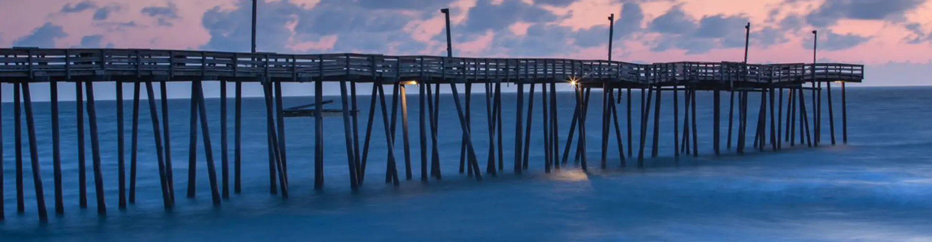 A long wooden fishing pier stretching out over calm blue water at dusk