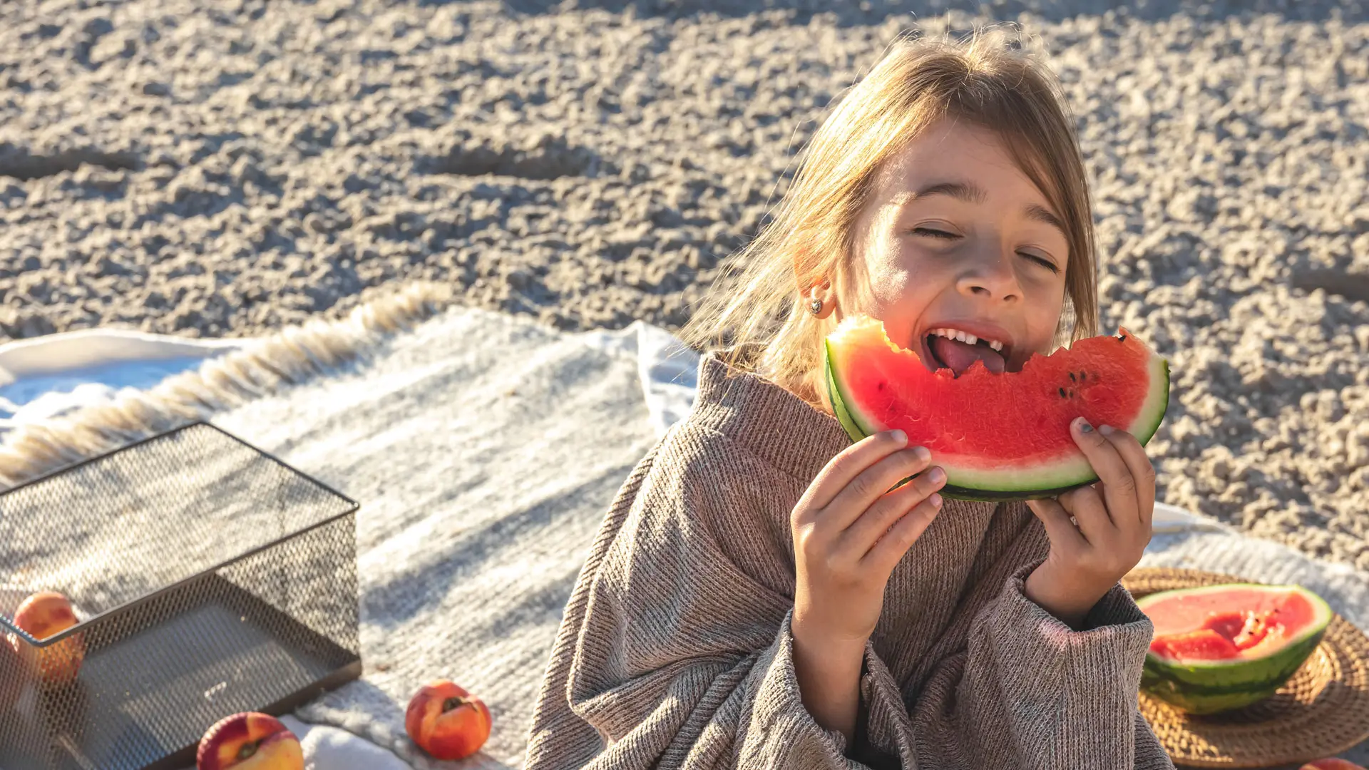 A girl wrapped in a blanket on the beach, eating a large slice of watermelon