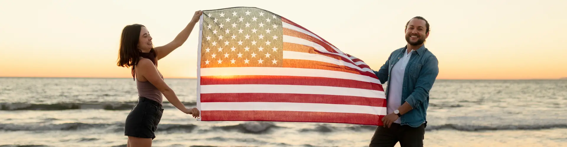 A couple holding an American flag stretched open between them on the beach at sunset