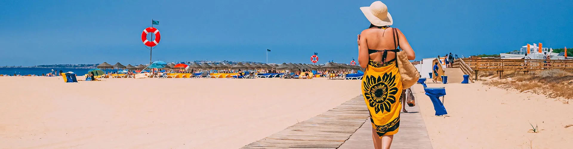 A woman in a wide sun hat walking a boardwalk down to a busy beach of umbrellas