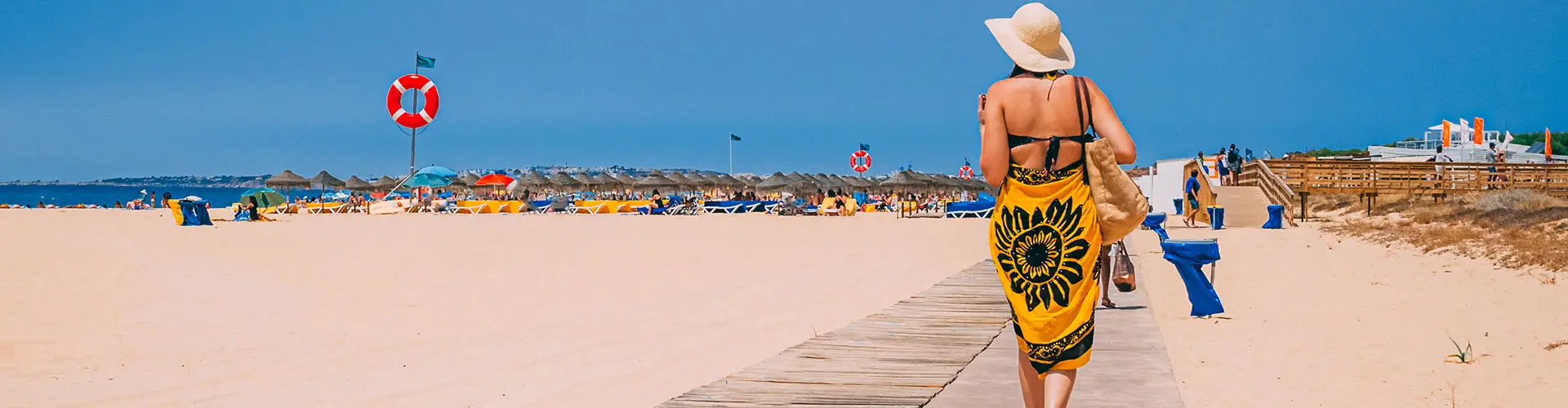 A woman in a wide sun hat walking a boardwalk down to a busy beach of umbrellas