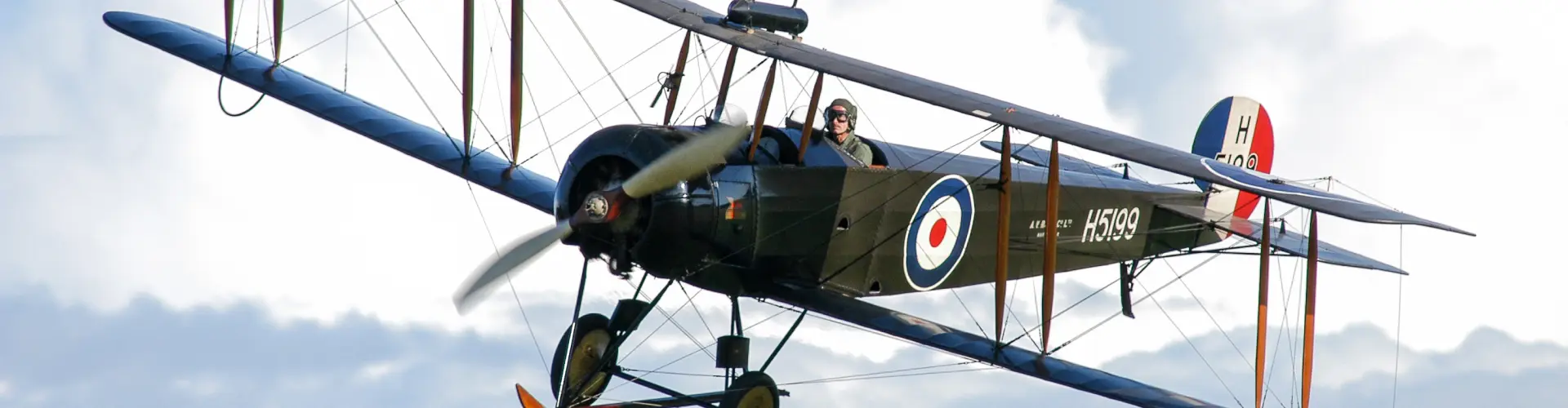 A vintage biplane in flight, its open cockpit and roundel markings visible against cloud