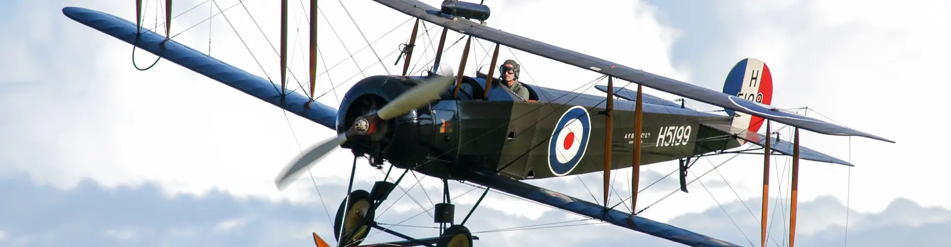 A vintage biplane in flight, its open cockpit and roundel markings visible against cloud